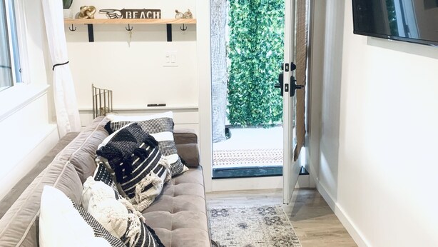 Ein modernes Wohnzimmer mit einer Couch, einem Flachbildfernseher, einer Holzregal mit Dekoration und Blick auf einen grünen Garten durch eine geöffnete Tür.