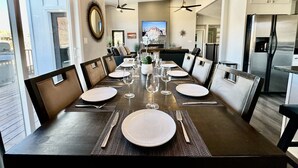 A dining area with a long table set for six, surrounded by chairs, with a kitchen in the background.