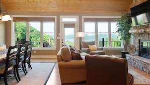 Un salon chaleureux avec une cheminée en pierre, un plafond en bois et de grandes fenêtres donnant sur un paysage pittoresque.