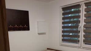 Een eenpersoonsbed met geruit beddengoed, een lichtschakelaar aan de muur en een raam met blinds.