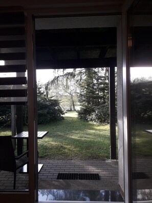 A view through a glass door into a garden with a brick pathway, a metal grate, and a bench. - MEIN HAVELURLAUB: Holiday home "HAVELIDYLL" Directly on the water! Nature with a vision (Oranienburg)