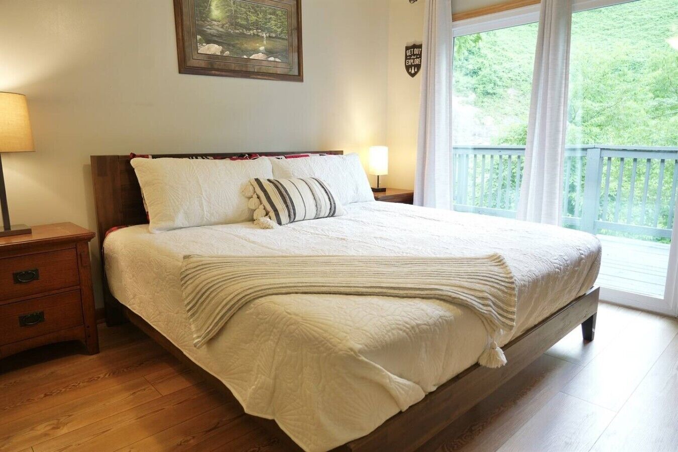 Un dormitorio con cama de madera, mesita de noche, lámpara, un cuadro enmarcado en la pared y balcón con vistas a árboles.