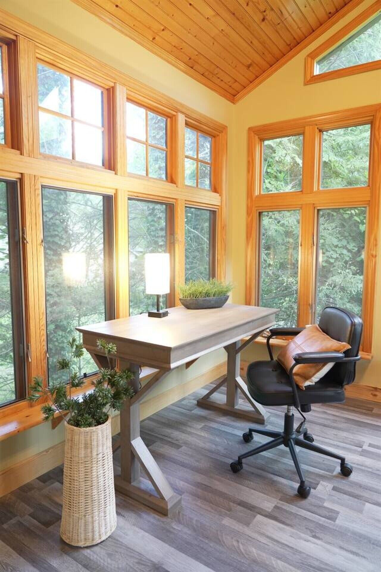A room with a wooden desk, a chair, a lamp, and a large window with a view of trees.