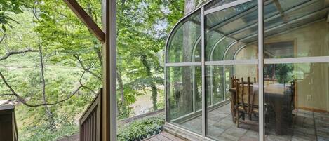 A covered outdoor area with a glass enclosure, wooden deck, and a view of a house with a dining table and chairs.
