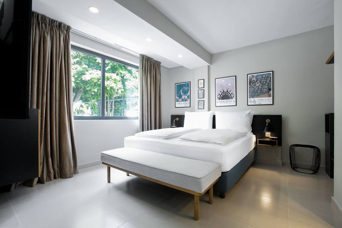 Quarto moderno com uma cama grande, um banco e vista para o verde lá fora.