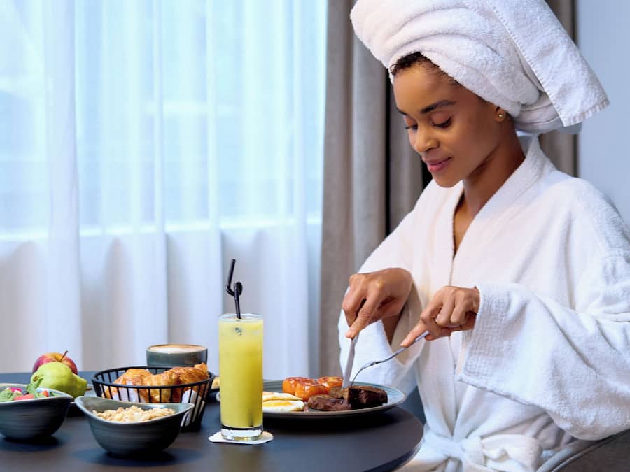 A person in a white bathrobe with a towel on their head, sitting at a round table with breakfast items including toast, fruit, and a glass of juice.