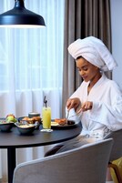 A person in a white bathrobe with a towel on their head, sitting at a round table with breakfast items including toast, fruit, and a glass of juice.