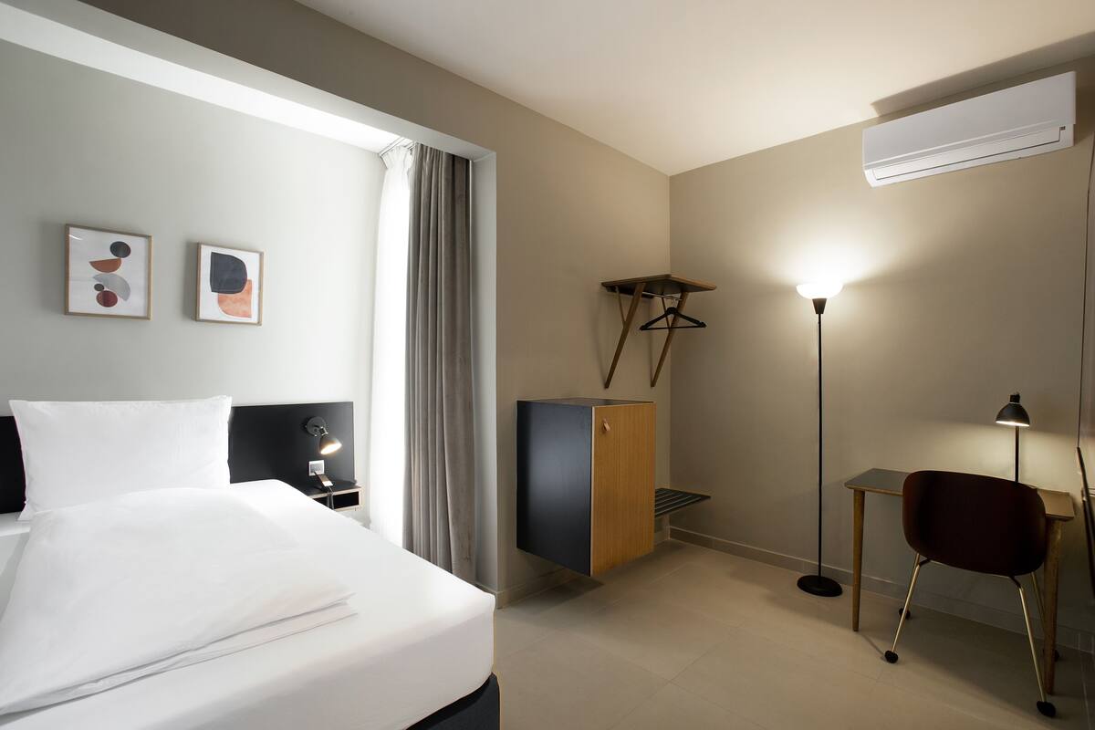 Quarto de hotel com cama, escrivaninha, guarda-roupa e duas fotografias emolduradas na parede.