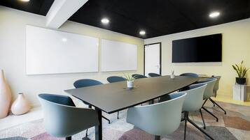 A conference room with a long table, six chairs, whiteboards, a TV, and a potted plant.