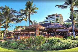 Un resort con estructuras de techo de paja, palmeras y un edificio de varios pisos al fondo.