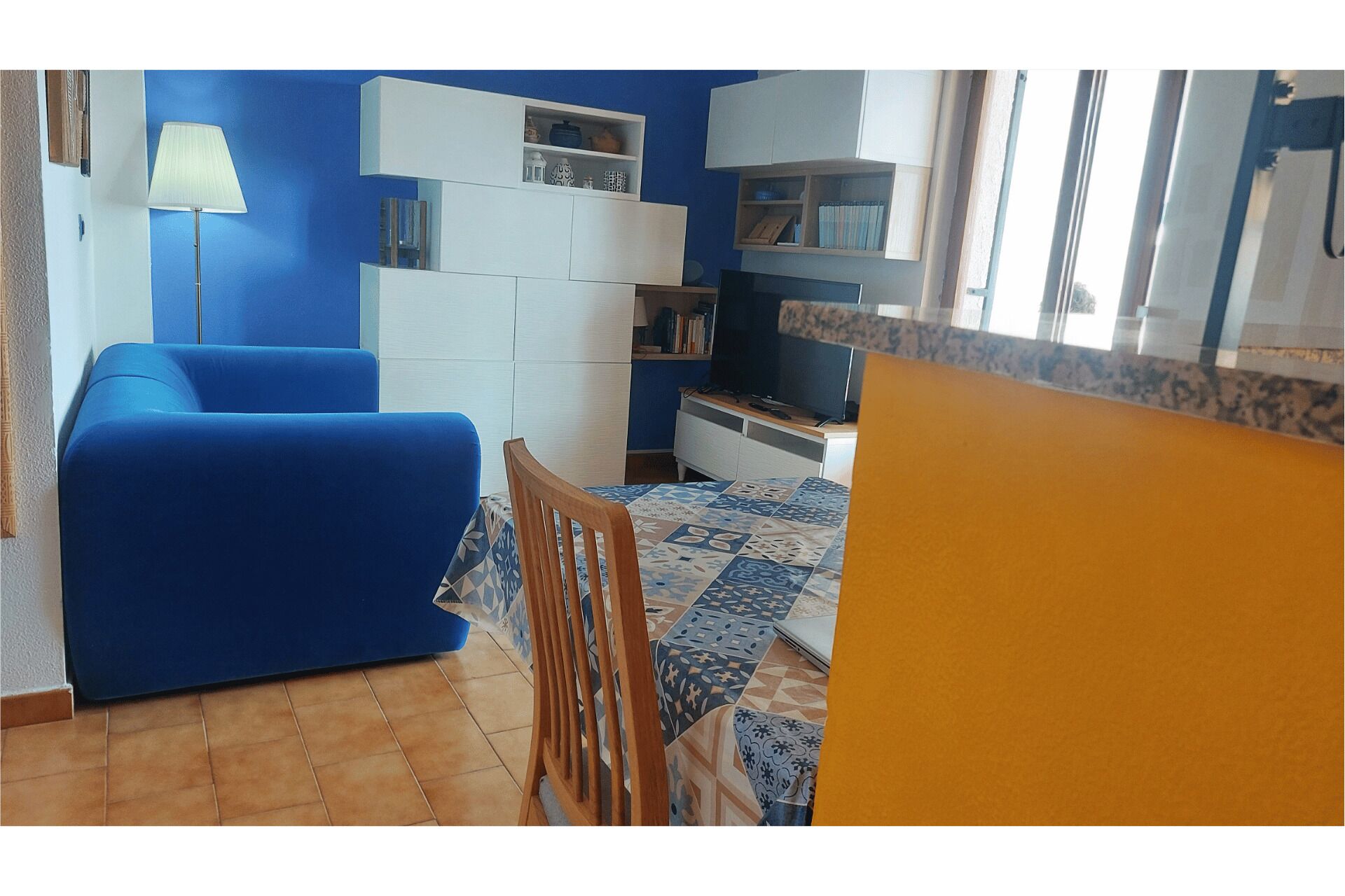 Uma sala de estar com um sofá azul, uma cadeira de madeira, uma televisão e uma estante de livros.