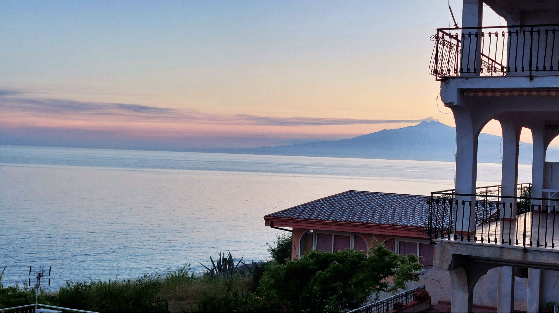 A coastal building with a balcony overlooking a calm sea and a distant mountain.