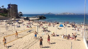 Een druk strand met een volleybalnet op de voorgrond en een rotsklif in de verte.