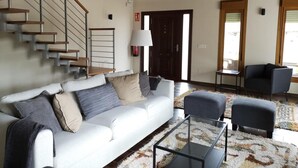 Un salón espacioso con un sofá blanco, dos sillones, una mesa de centro de cristal y una escalera con escalones de madera.