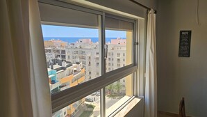 Ein Zimmer mit einem großen Fenster, das Blick auf eine Stadtlandschaft und das Meer gewährt. An der Wand ist ein Holzschrank mit einer Aufschrift zu den Kaffeeangeboten zu sehen.