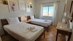 Ein Hotelzimmer mit zwei Einzelbetten, einem Nachttisch mit Lampe, einem Ventilator und einem Fenster mit Vorhängen.