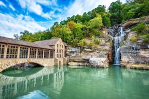 Eine Steinbrücke über ruhigem Wasser, ein rustikales Gebäude mit großen Fenstern und ein Wasserfall, der einen felsigen Klippen hinabfließt.