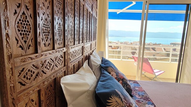 A room with a wooden carved headboard, a bed with floral-patterned bedding, and a view of the sea through large windows.