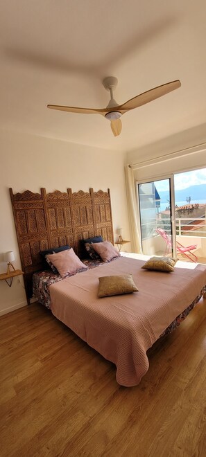 Un dormitorio con una cama grande, ventilador de techo, cabecera de madera y vista al mar a través de una ventana.