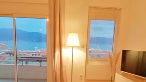 A modern hotel room with a laptop on a wooden desk, a TV on the wall, a lamp, and a view of a cityscape and body of water through large windows.