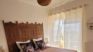 A bedroom with a wooden headboard, a large bed with floral bedding, a hanging light fixture, and a view through sheer curtains.
