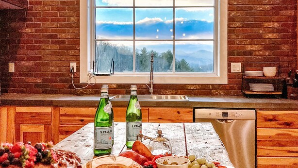 Una cocina con pared de ladrillo rústica, un ventanal con vistas, armarios de madera y una mesa de madera con comida y bebidas.