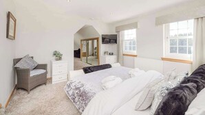 A spacious bedroom with a large bed, a sitting area with a chair, a dresser, and a television mounted on the wall. - Greenknowes Farmhouse with Hot Tub and Bar (Blairadam)