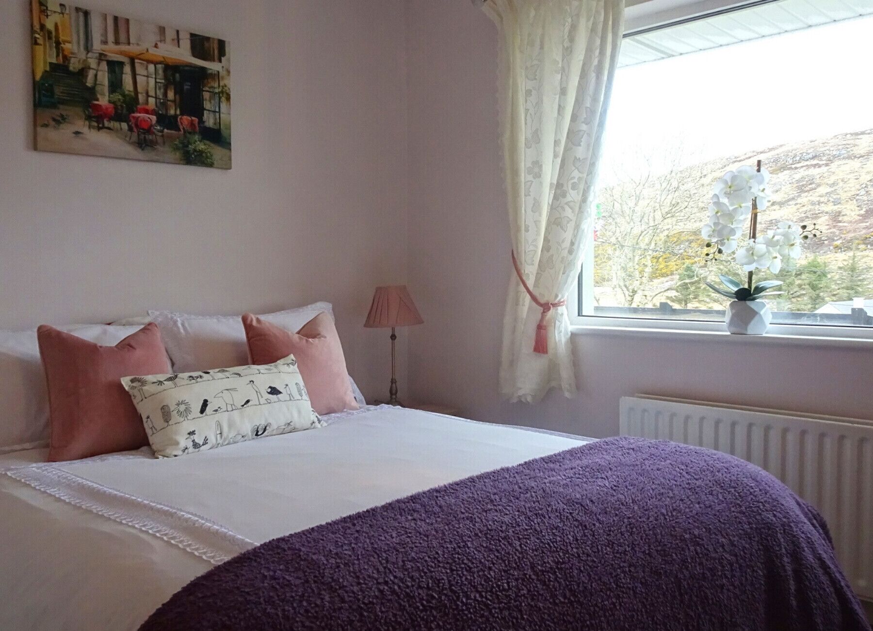 A bedroom with a bed, pillows, a window with curtains, a radiator, and a painting on the wall.