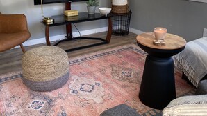 A living room with a flat-screen TV, a round coffee table, a woven ottoman, a patterned rug, and a window with blinds. - East Main Downtown Bungalow  (Ashland)