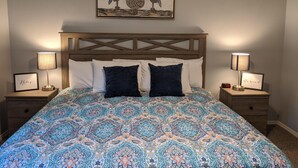 Une chambre à coucher avec une couvre-lit à motifs, une tête de lit en bois, deux lampes de chevet et des œuvres d’art encadrées au-dessus du lit.