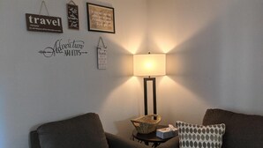 Un salon chaleureux avec un canapé gris, un coussin à motifs, une petite table sur laquelle se trouvent un panier et une boîte de mouchoirs, ainsi que des œuvres d’art murales inspirantes.