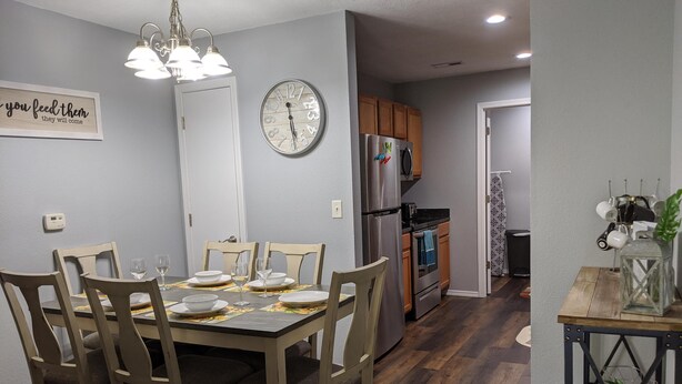 Un coin repas avec une table en bois dressée pour quatre personnes, une horloge au mur et une cuisine en arrière-plan.