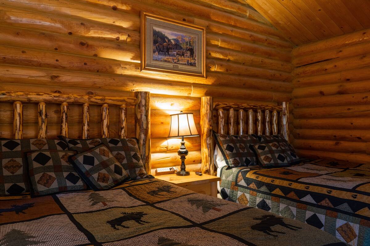 Una habitación tipo cabaña de troncos con dos camas, un cuadro en la pared y una lámpara en una mesita de noche.