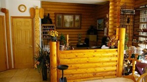 A log cabin interior with a wooden reception desk, a clock on the wall, and a variety of plants and decorations.