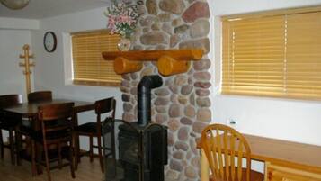Una habitación acogedora con pared de piedra, una mesa de madera, sillas, chimenea y ventana con persianas.