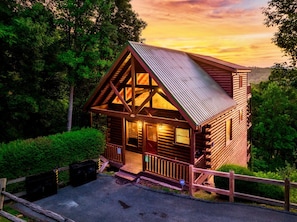 A wooden cabin with a sloped roof, surrounded by dense greenery, and accessible by a paved path.