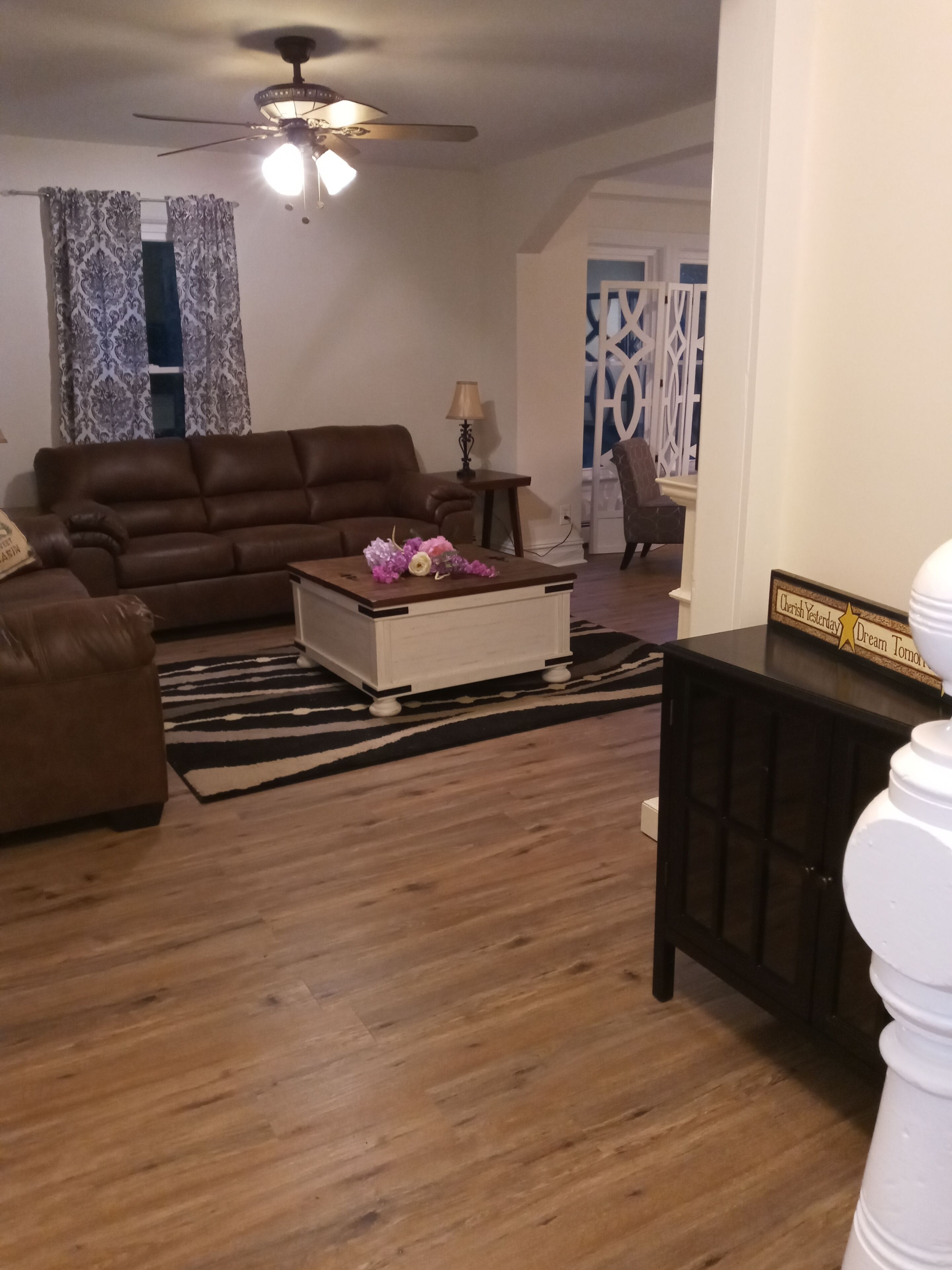 A living room with a brown sofa, a white coffee table with flowers, a ceiling fan, and a zebra-patterned rug.
