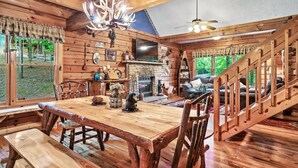 L’intérieur d’un chalet rustique comprend une table à manger en bois, des bancs, une cheminée, un téléviseur et un escalier.