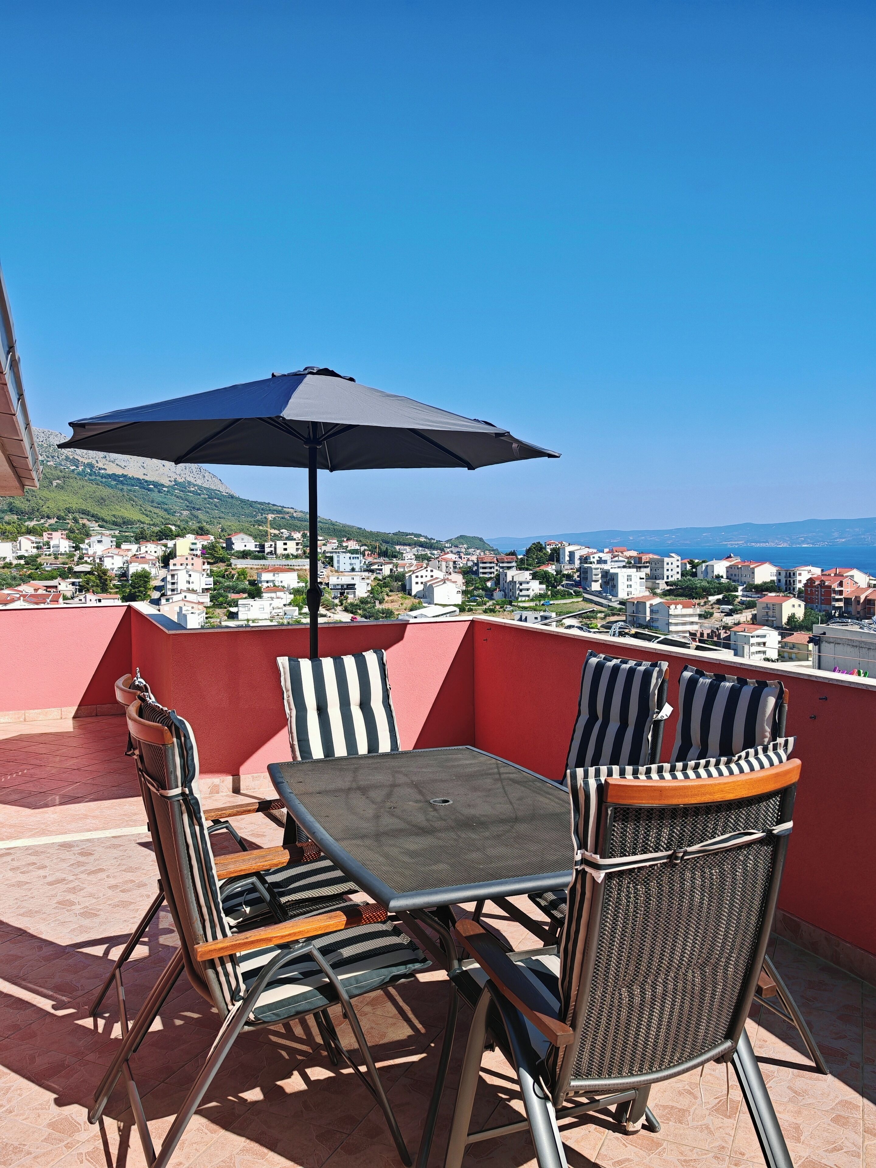 A terrace with a table and chairs, an umbrella, and a view of a coastal town.