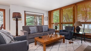 A modern living room with a grey sofa, a wooden coffee table, and a blue armchair. There are two lamps on side tables, and a large window with wooden blinds.