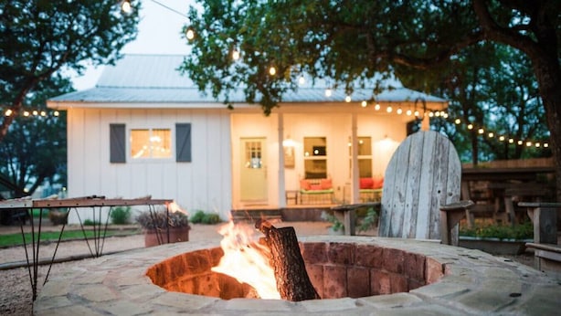 A stone fire pit with a fire burning, surrounded by a stone bench and a wooden table. In the background, there is a white building with a porch and string lights.