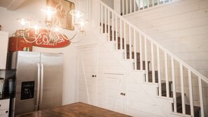 A kitchen with a stainless steel refrigerator, a wooden dining table, white wooden stairs, and a chandelier.