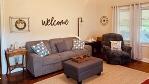 A living room with a gray sectional sofa, a dark armchair, a wooden side table, and a wall-mounted clock. - Cozy, Country Cottage close to Oaklawn, near Bypass (Hot Springs)