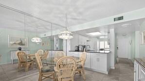 A modern kitchen with white cabinets, a central island, and a dining area with wicker chairs and a glass table. - Gulf Coast / Direct Beachfront View - Steps to Sand & Sunset (Fort Myers Beach)