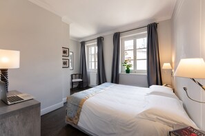 A hotel room with a bed, a desk with a laptop, a chair, a window with curtains, and a nightstand with a lamp. - Goldoni  Novella Suite B2 (Firenze)