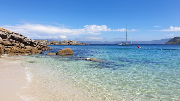 A beach with clear turquoise water, rocky shoreline, and a sailboat in the distance.