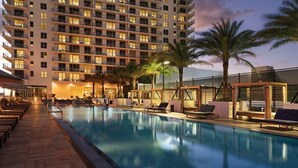 Area piscina di un hotel con una grande vasca, lettini prendisole e un grattacielo sullo sfondo.