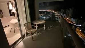 Un balcone con tavolo e sedie, vista sulla città di notte e scorcio dell'interno che include un divano e una lampada.