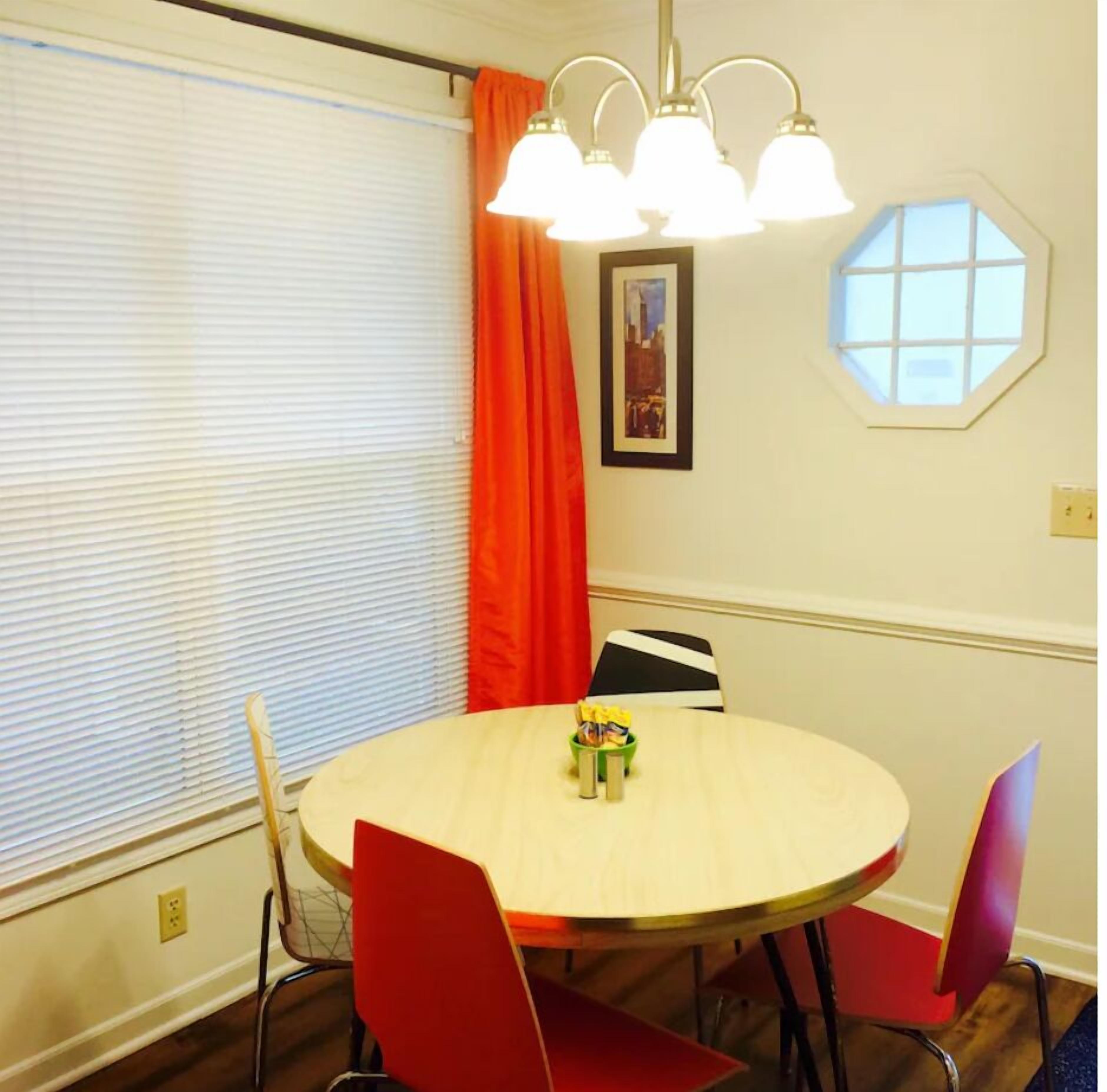 Een eethoek met een ronde tafel, rode stoelen, een raam met jaloezieën, een rood gordijn en een wandlamp.