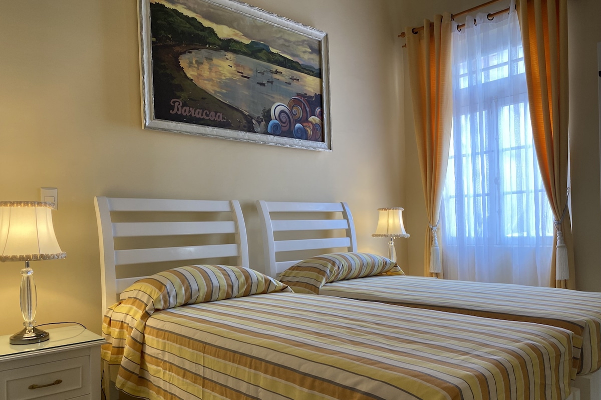 A double bed with striped bedding, a nightstand with a lamp, a framed picture on the wall, and a window with curtains.